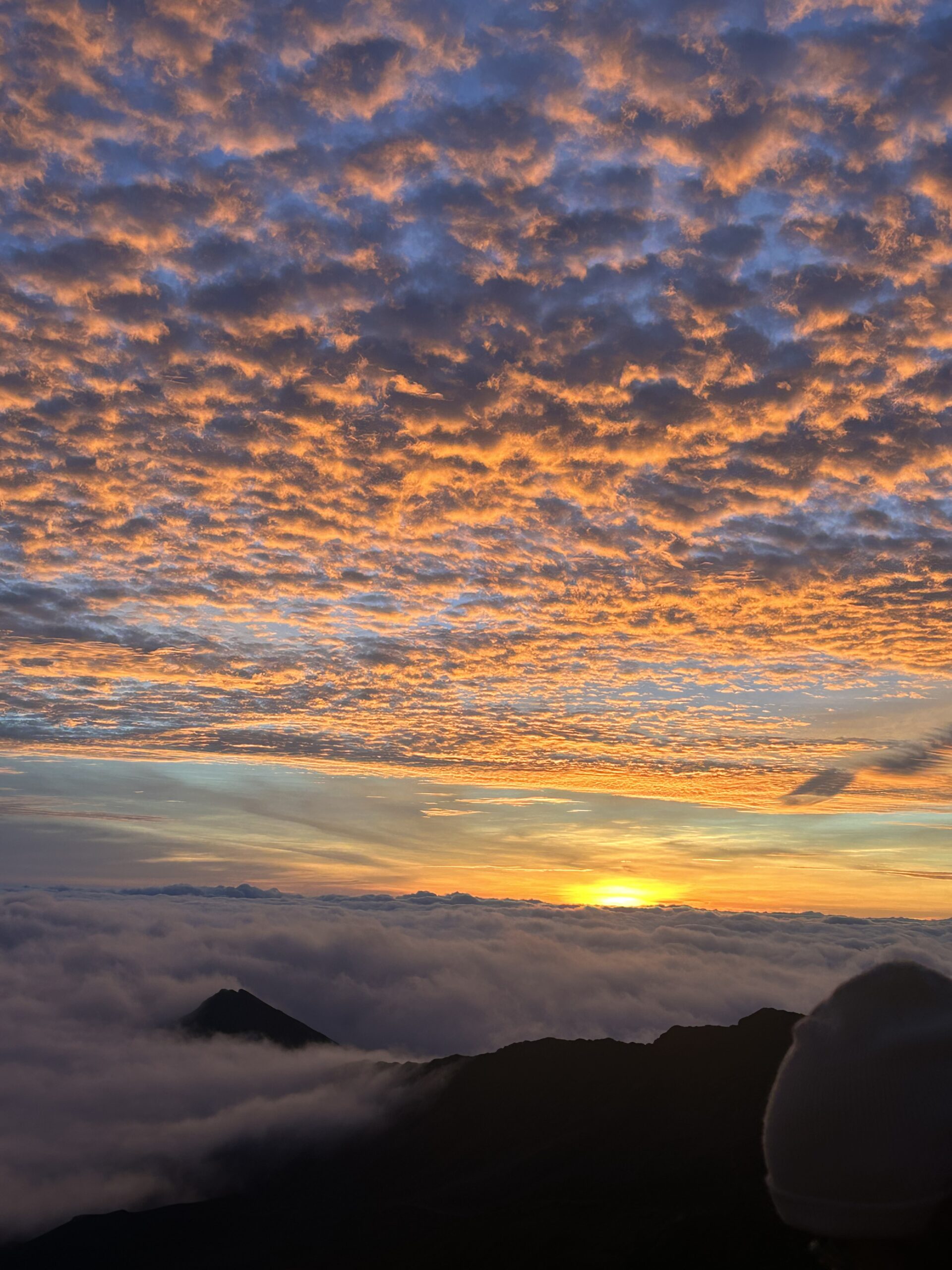 Vibrant sunset over a sea of clouds, with golden hues illuminating the sky and silhouetting distant mountains—a breathtaking natural scene.