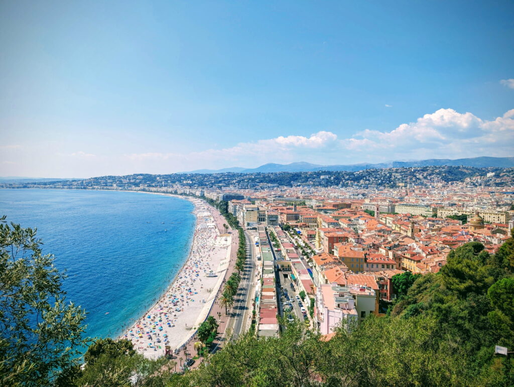 Panoramic view of Nice's coastline, featuring a vibrant beach, clear blue waters, and the city's colorful buildings under a sunny sky.