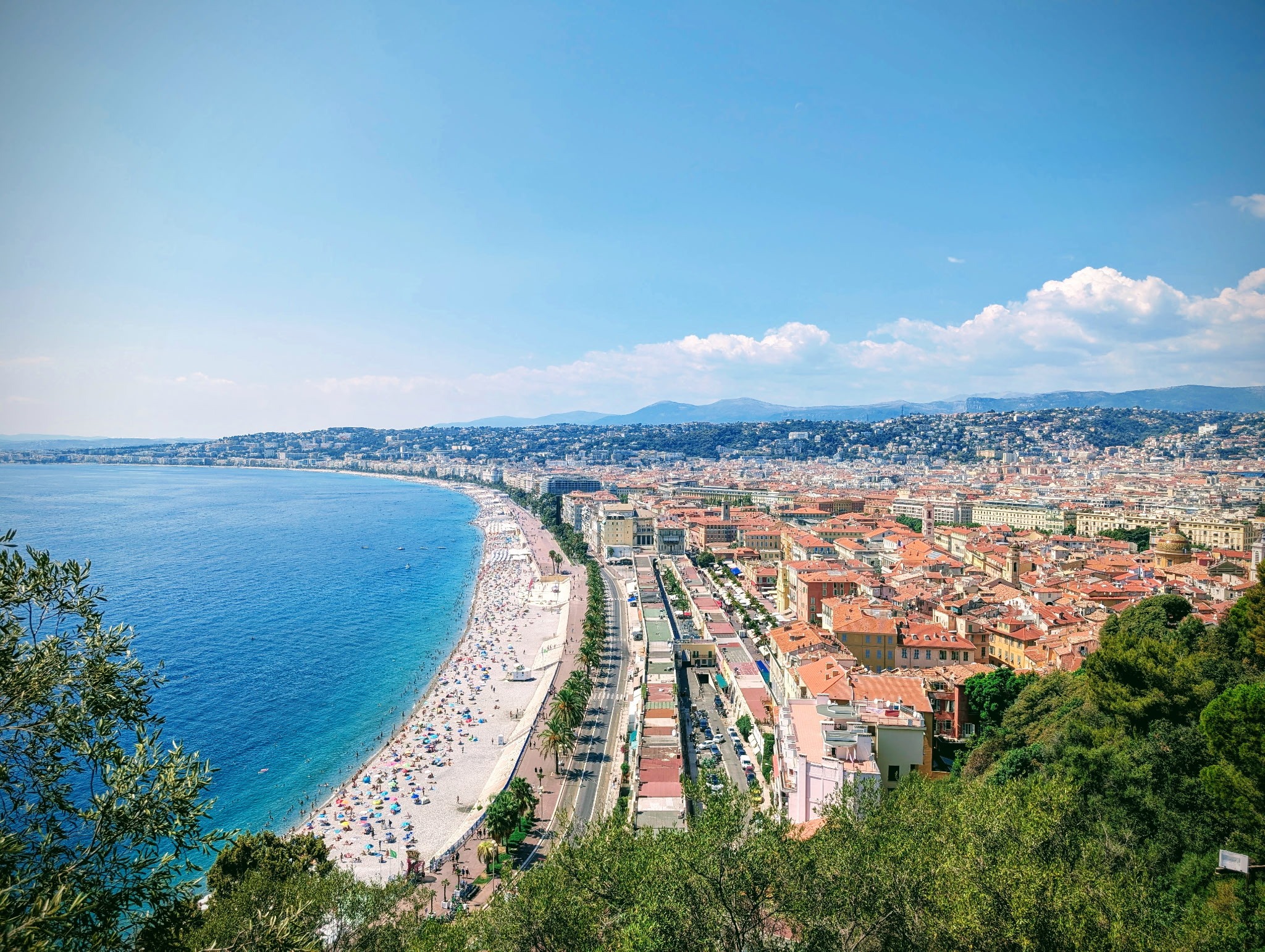 Panoramic view of Nice's coastline, featuring a vibrant beach, clear blue waters, and the city's colorful buildings under a sunny sky.
