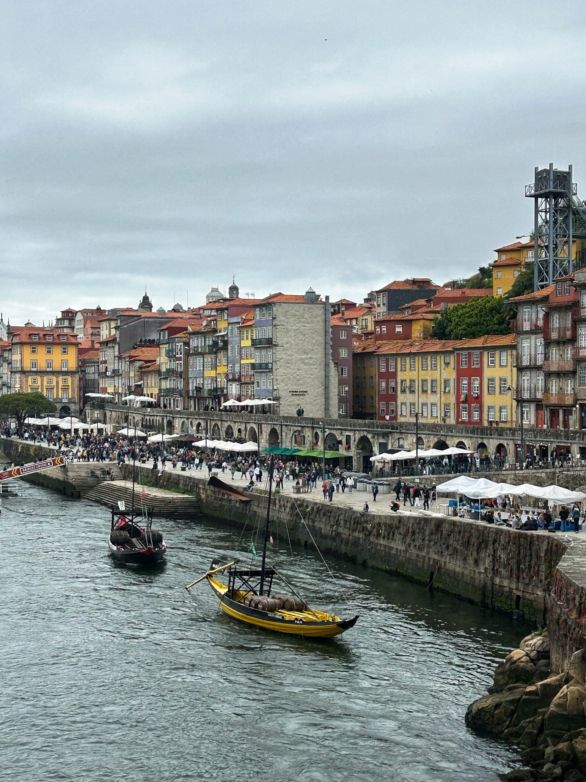 Colorful buildings line the riverside in Porto, Portugal, where traditional boats navigate the water and people stroll along the promenade.