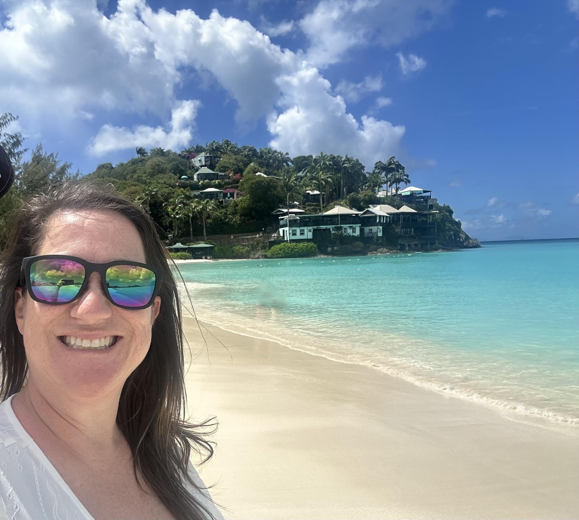 A woman with long hair smiles on a beach with turquoise water and a lush hillside in the background, showcasing a tropical vacation setting.