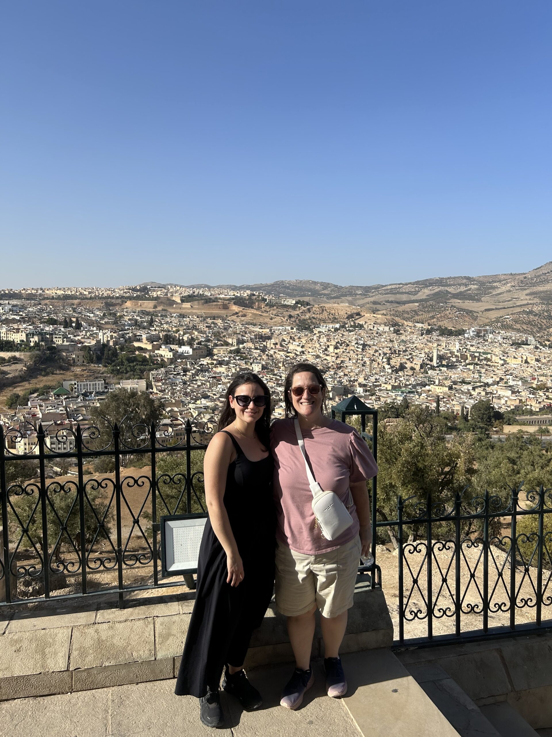 Two women pose together at a scenic viewpoint overlooking a sprawling city and hills, enjoying a sunny day.
